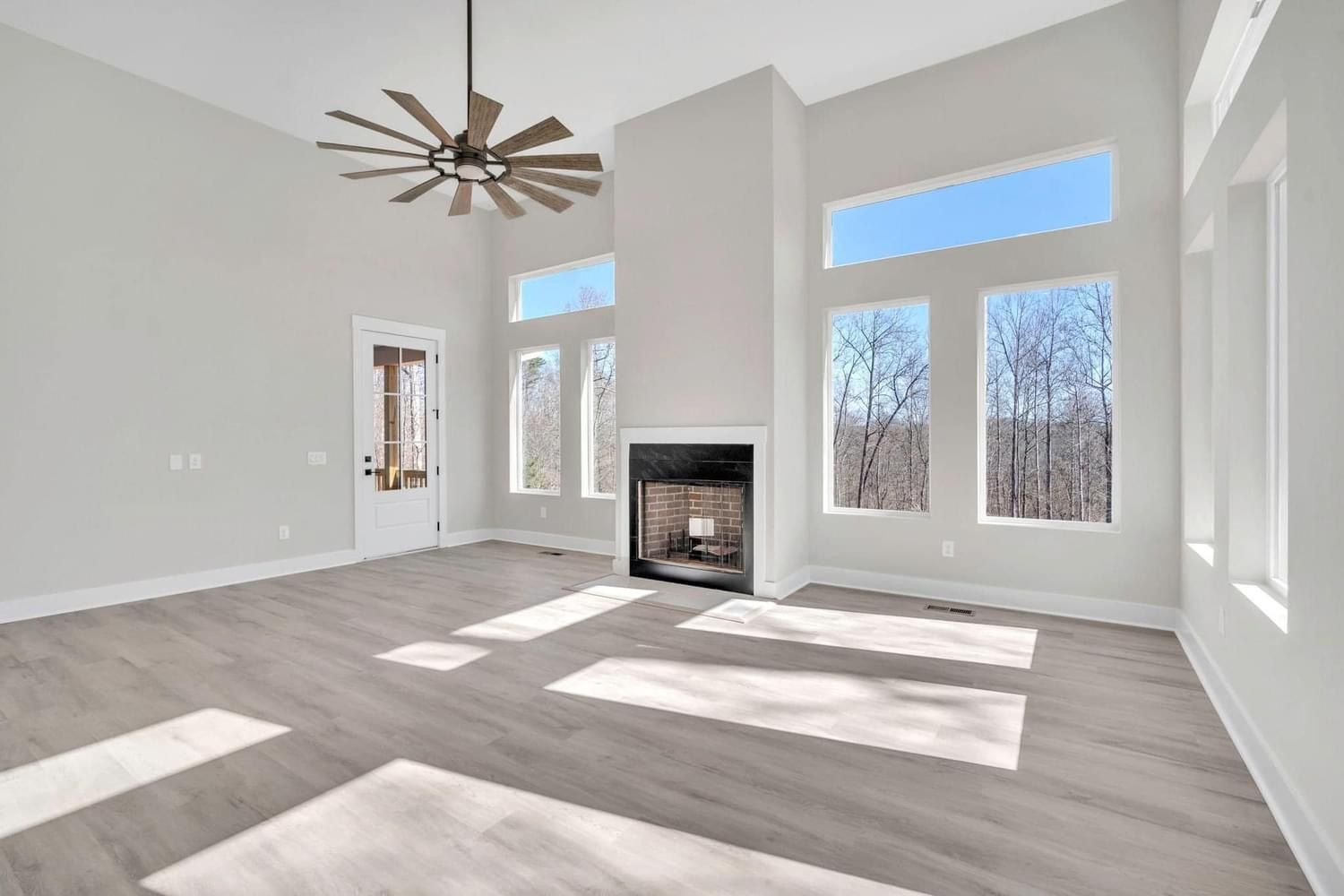 Bright empty living room with a fireplace, large windows, and sunlight on light wood floors