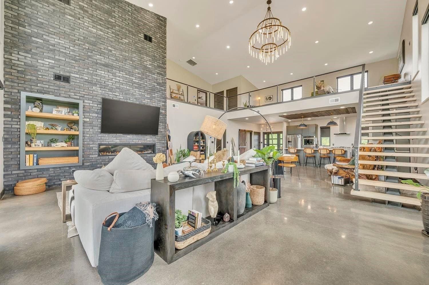 Bright modern living room with stone fireplace, staircase, large windows, and chandeliers