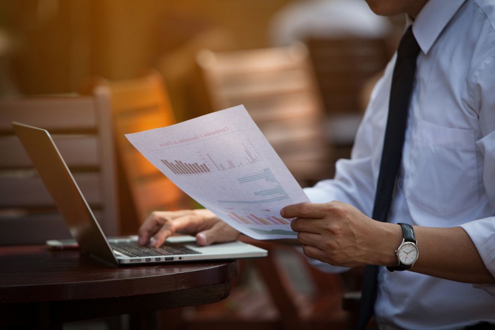 A Man Is Sitting at A Table Using a Laptop and Holding a Piece of Paper — Adding Value CFO Pty Ltd in Palmerston, NT