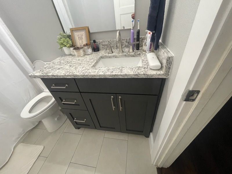 Small bathroom vanity with gray granite countertop, dark cabinets, sink, mirror, and toilet beside it.