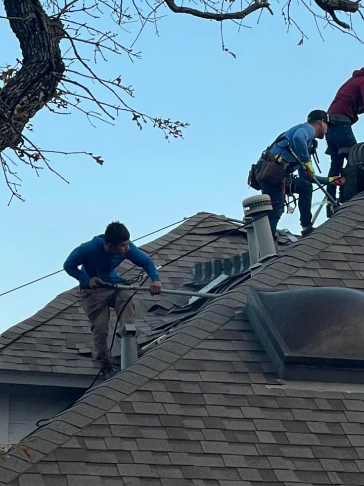Three roofers install equipment on a shingle roof. One pulls a wire while others work near skylight, blue sky.