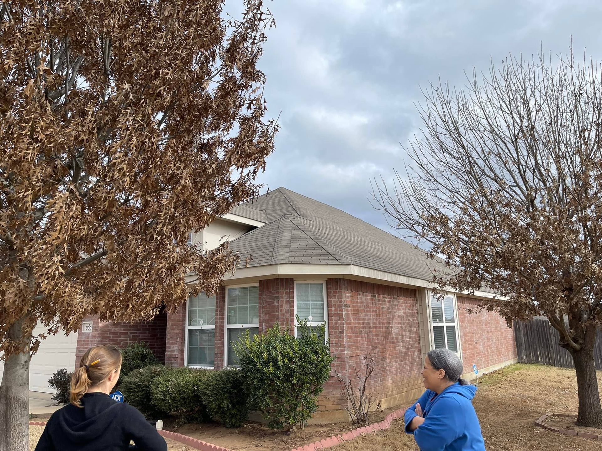 Two women in front of a brick house with brown trees and cloudy sky.