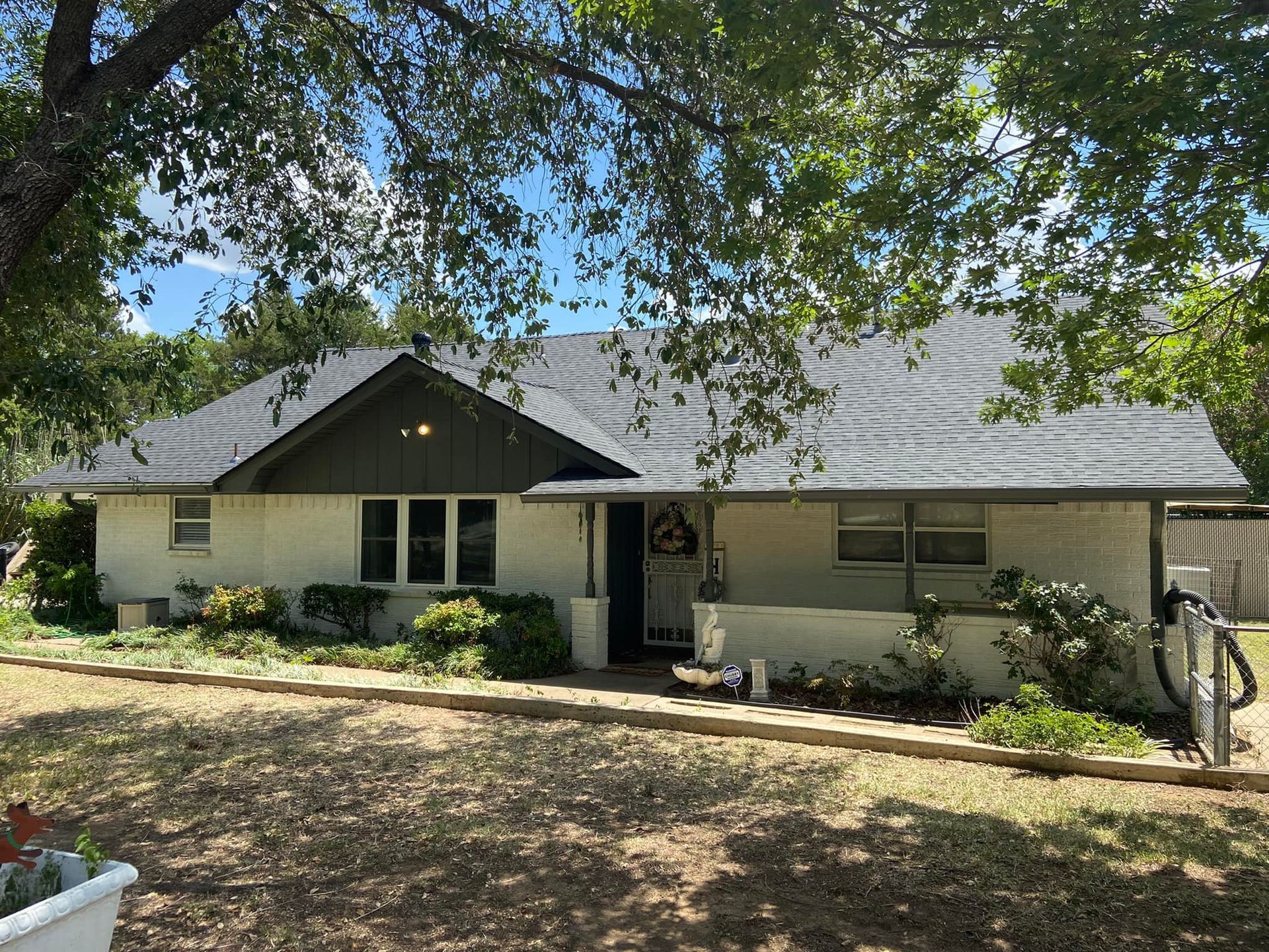 Single-story brick house with gray roof and porch, surrounded by trees and greenery under a blue sky.