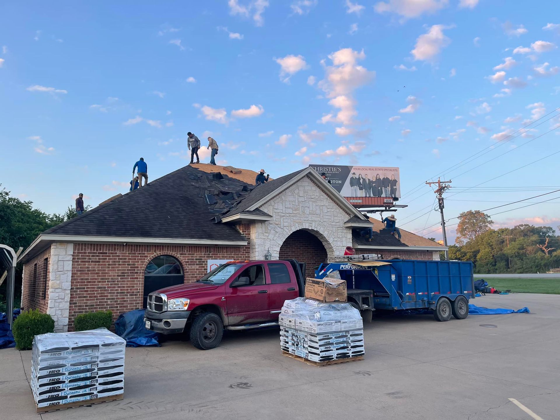 Roofing crew working on a building's roof. Red truck, trailer, shingles, and blue sky.
