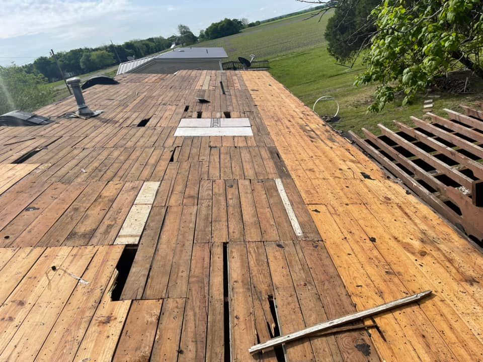 Wooden rooftop with planks removed, revealing gaps. Tools and surrounding green hillside are also visible.