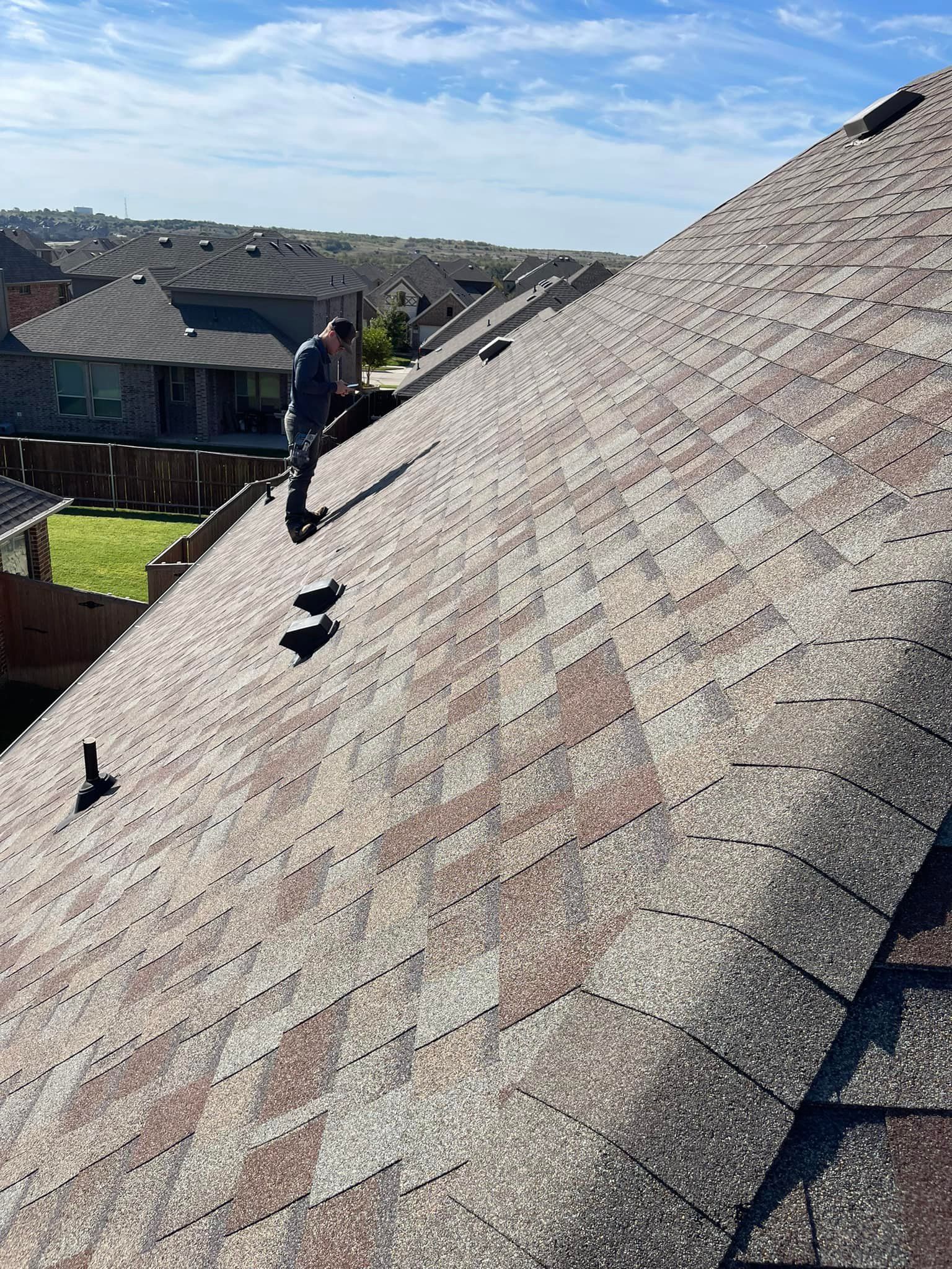 Roofer on a residential roof, inspecting shingles under a blue sky.