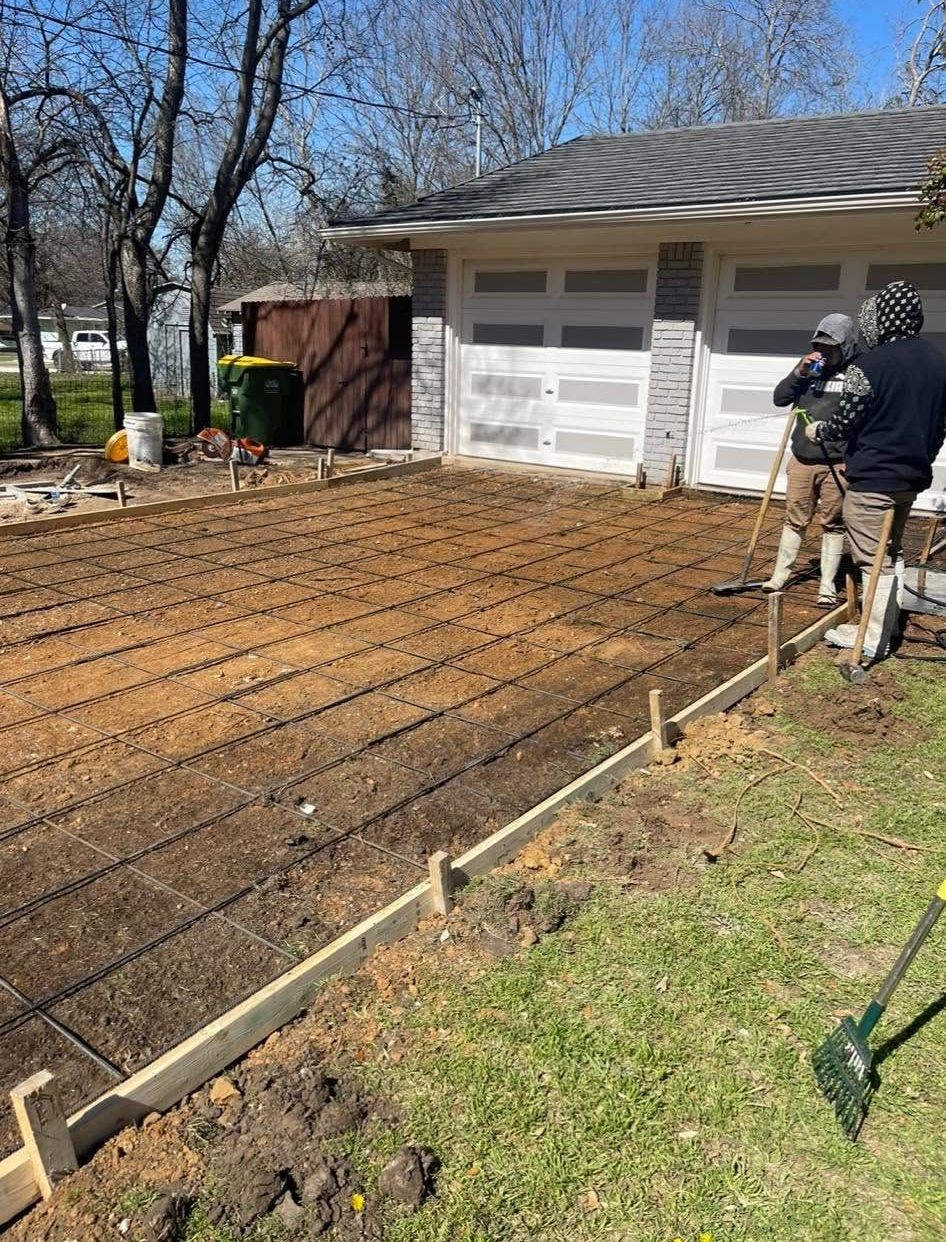 Two workers preparing a driveway for concrete. They're raking soil and setting wooden forms, near a garage and lawn.