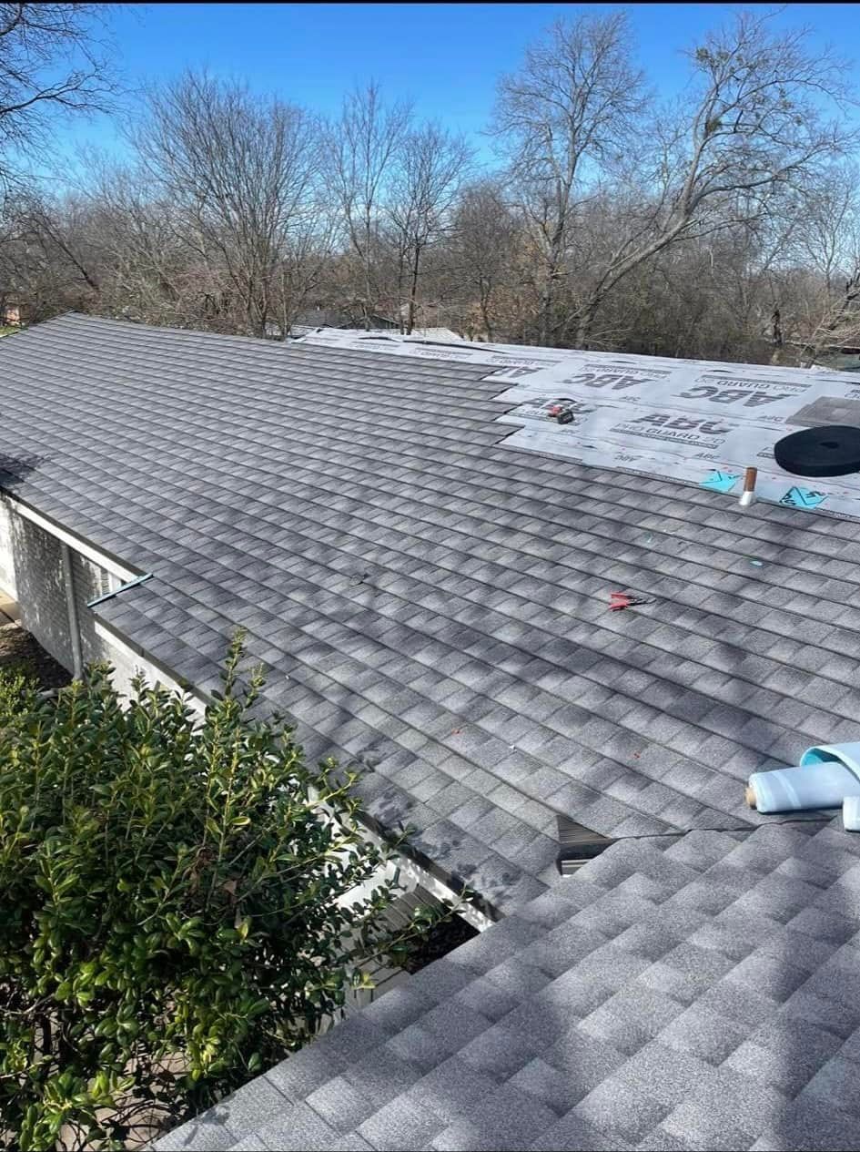 Gray shingle roof partially replaced on a residential building. Blue sky and trees in the background.