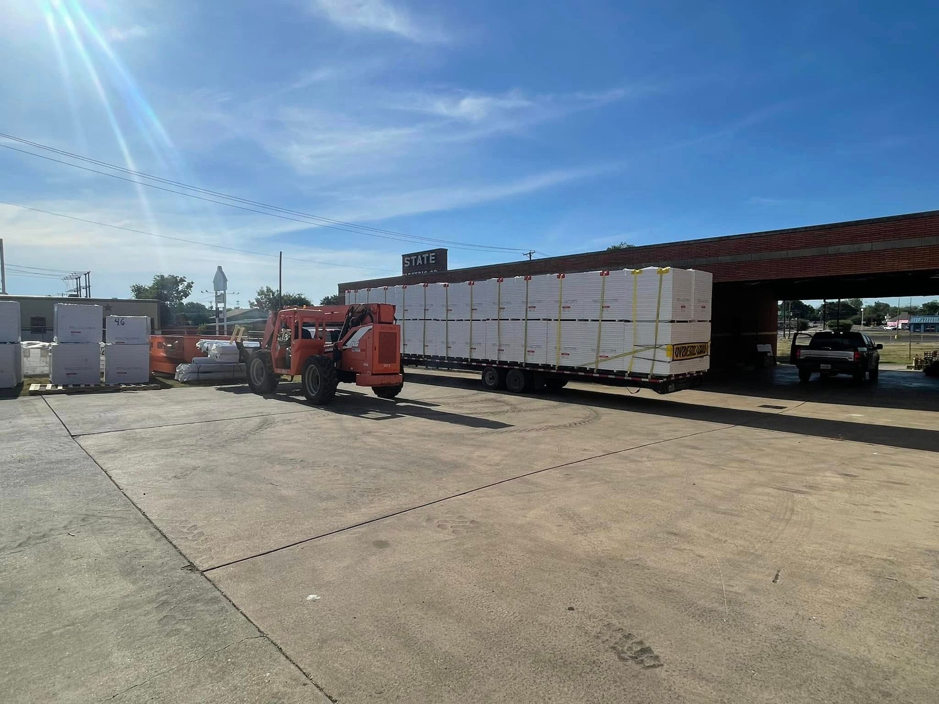 An orange forklift unloads boxes from a trailer in a loading area on a sunny day.