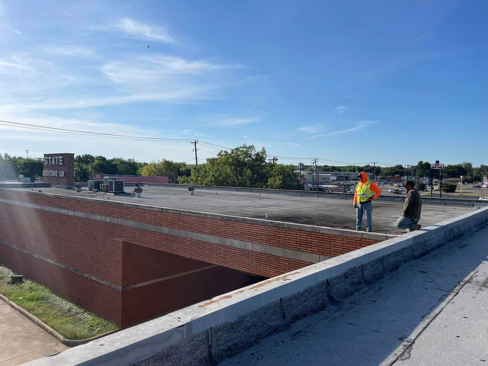 Two workers on a flat roof. One wears an orange vest. Brick wall in foreground, city background.