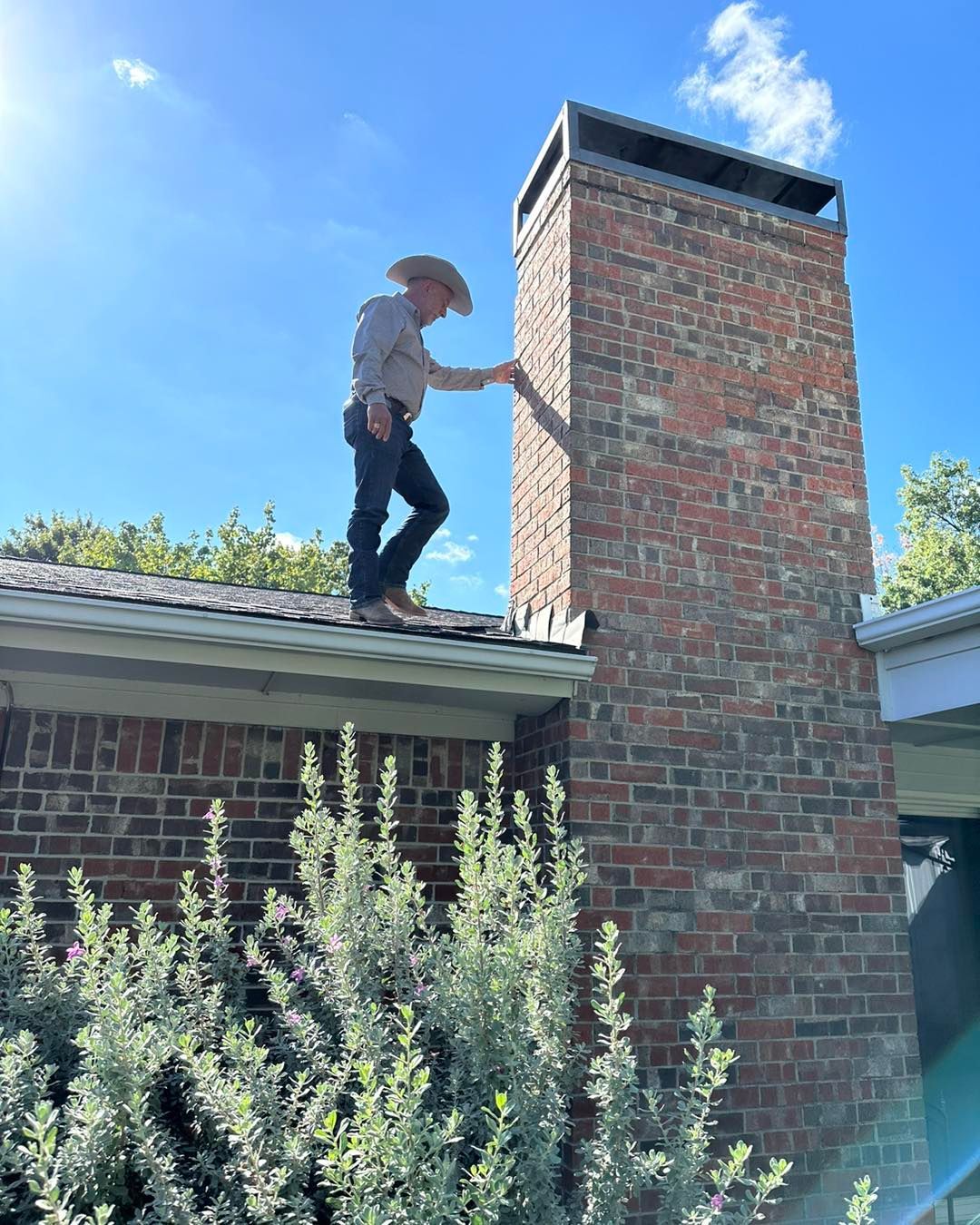 Man in cowboy hat on a roof, reaching toward a tall brick chimney. Bright sunny day.