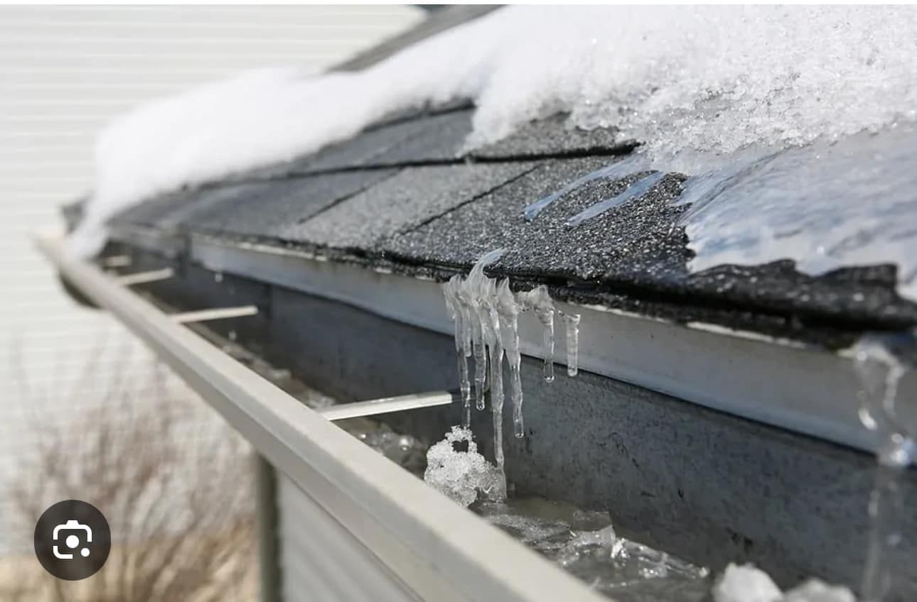 Snow and ice on a roof's edge, partially blocking a gutter. Ice is forming and dripping down.