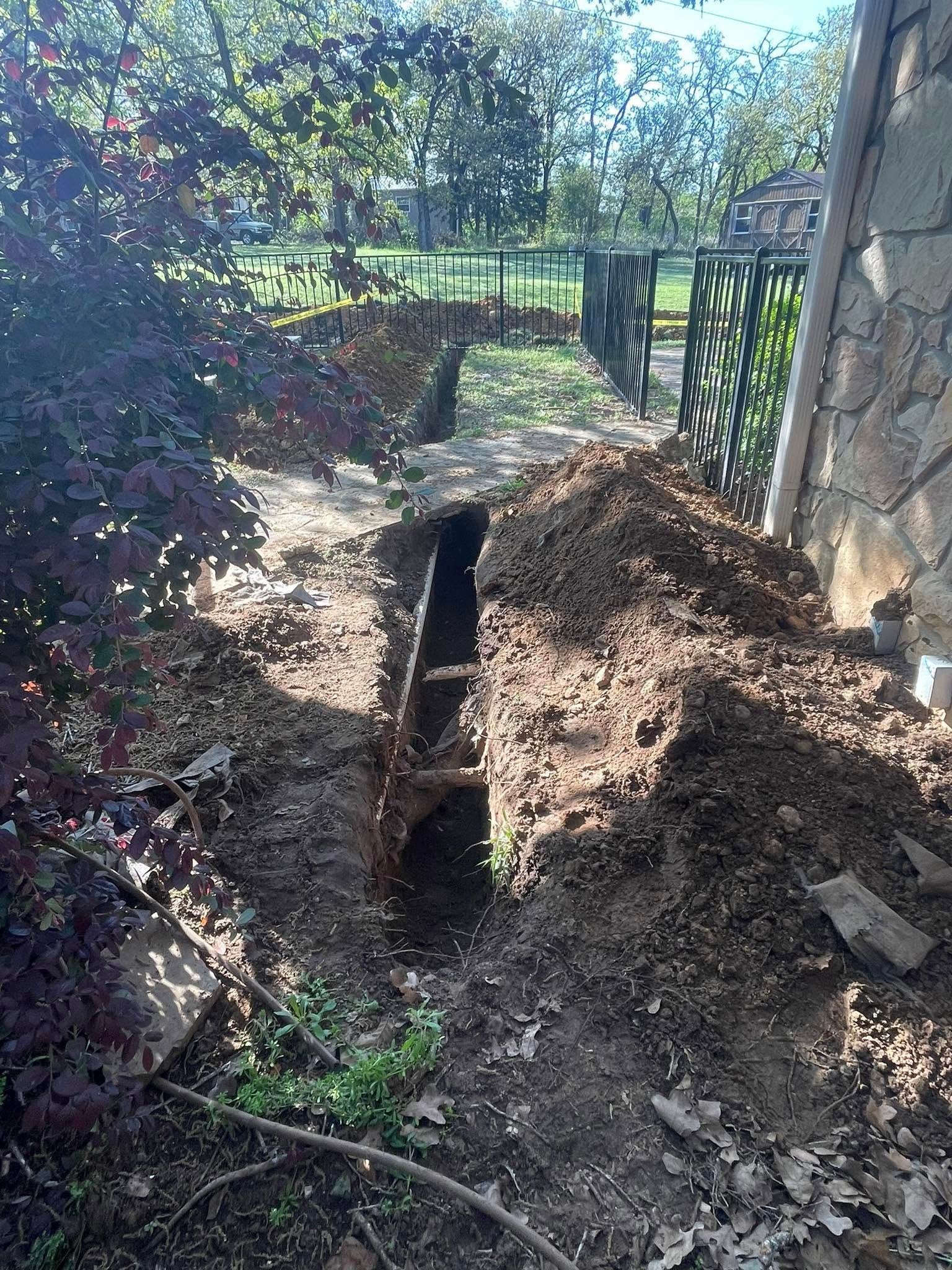 A trench dug along a fence, soil pile in foreground, trees and building in the background.