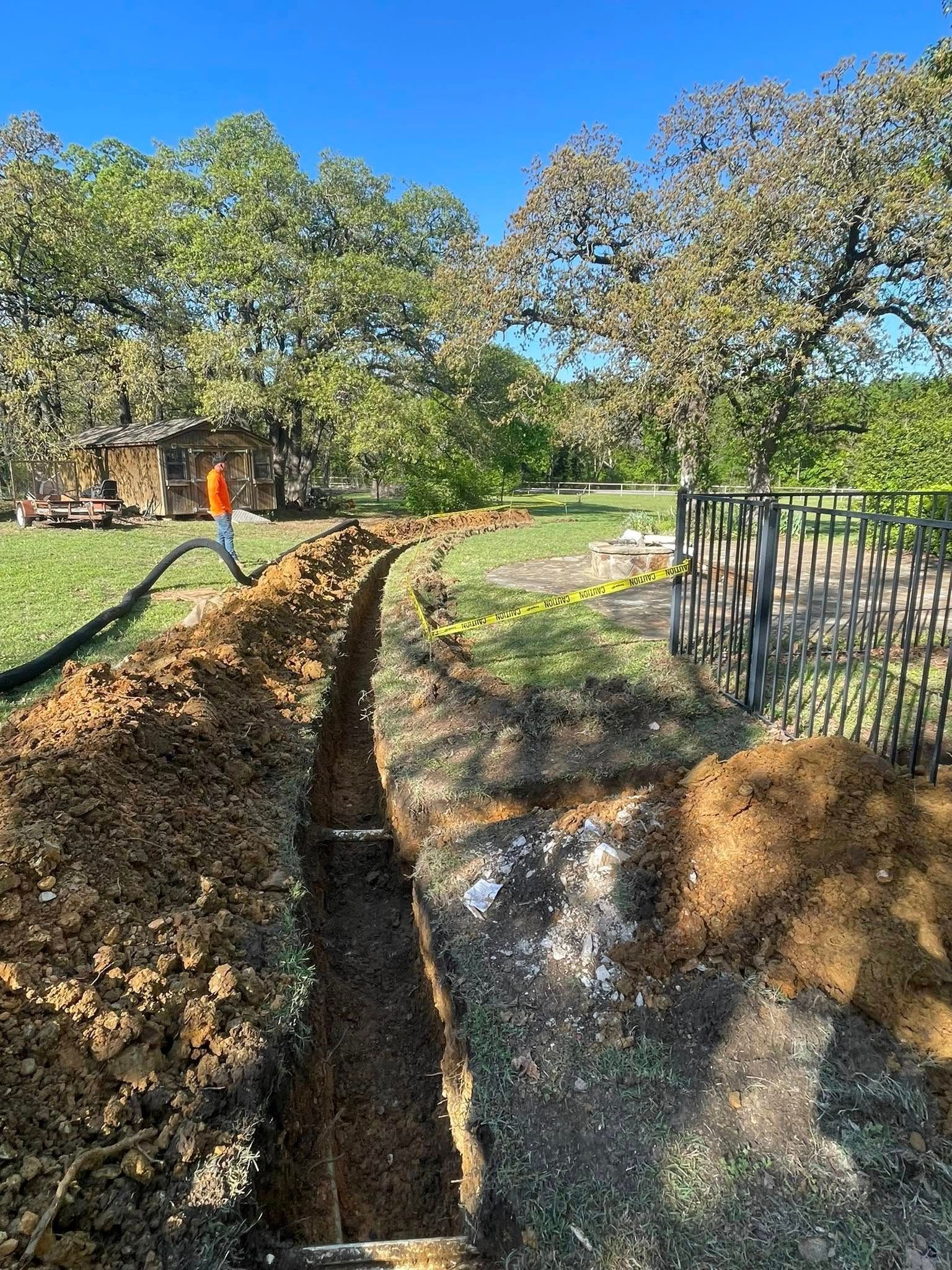 A trench dug in a grassy yard; a person stands in the distance near a building and fence.