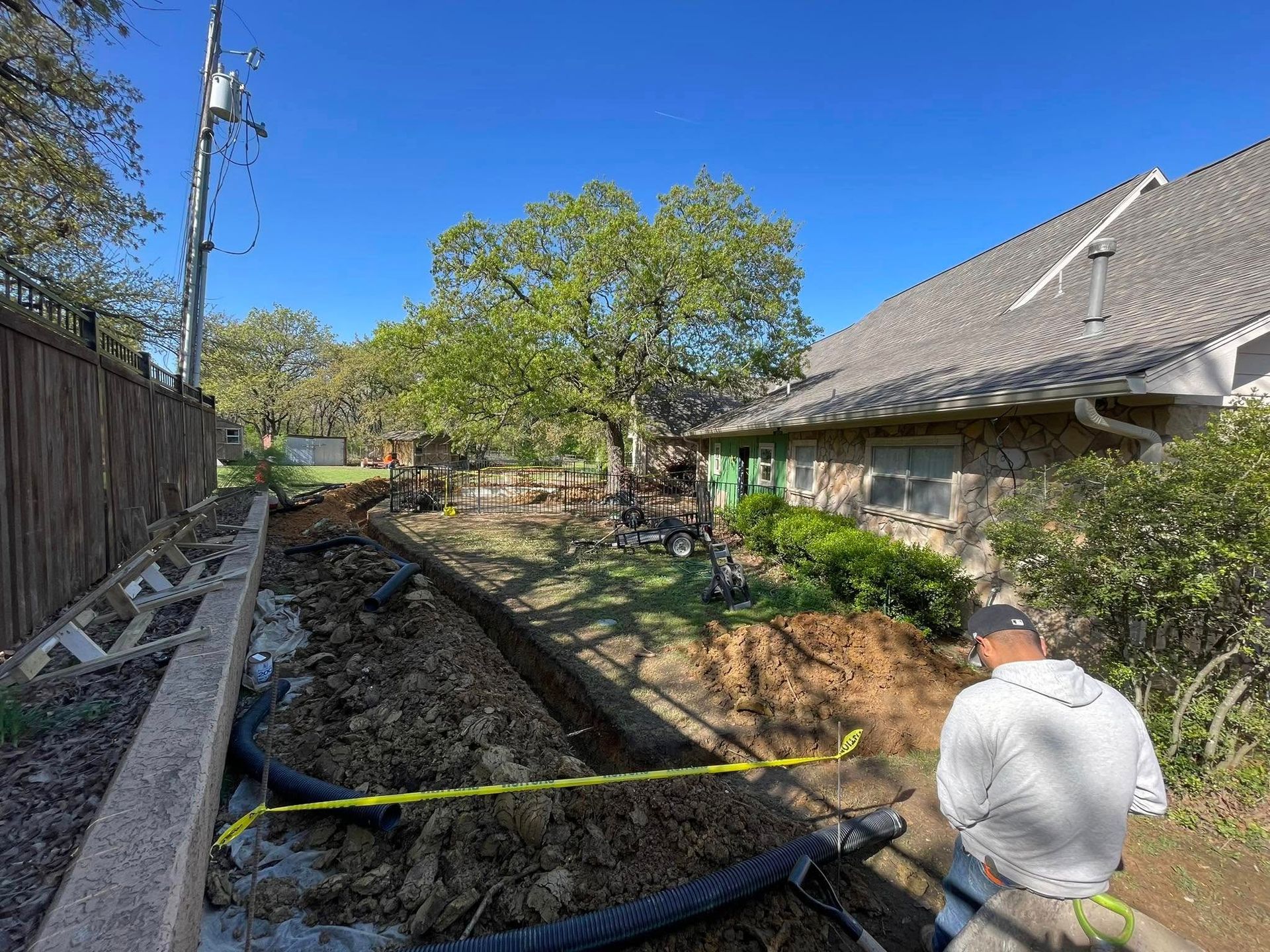 Man working on a trench next to a house and fence, with yellow caution tape and blue sky.