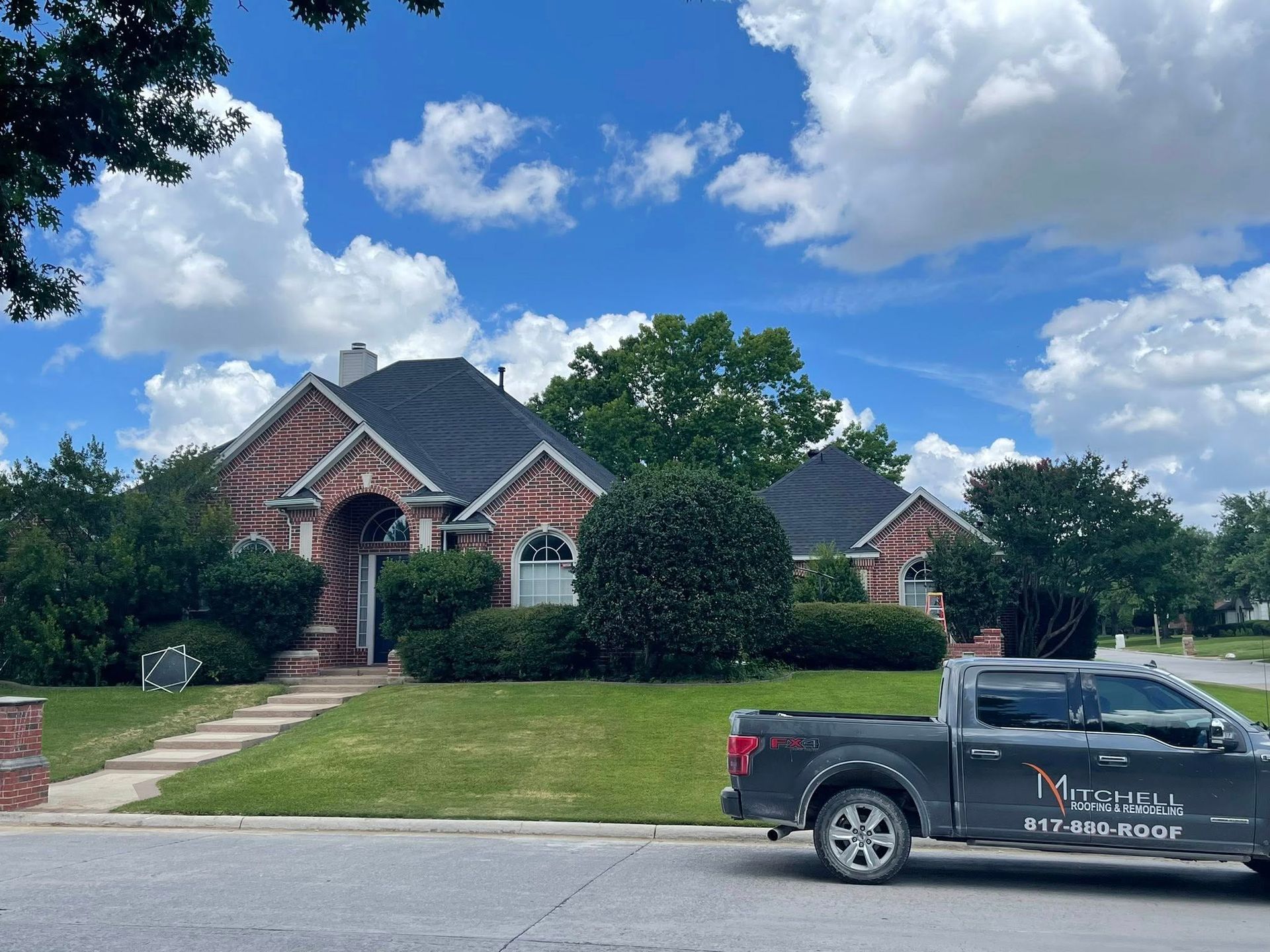 A brick house with a dark roof, a gray truck parked on the street, and a blue sky with clouds.