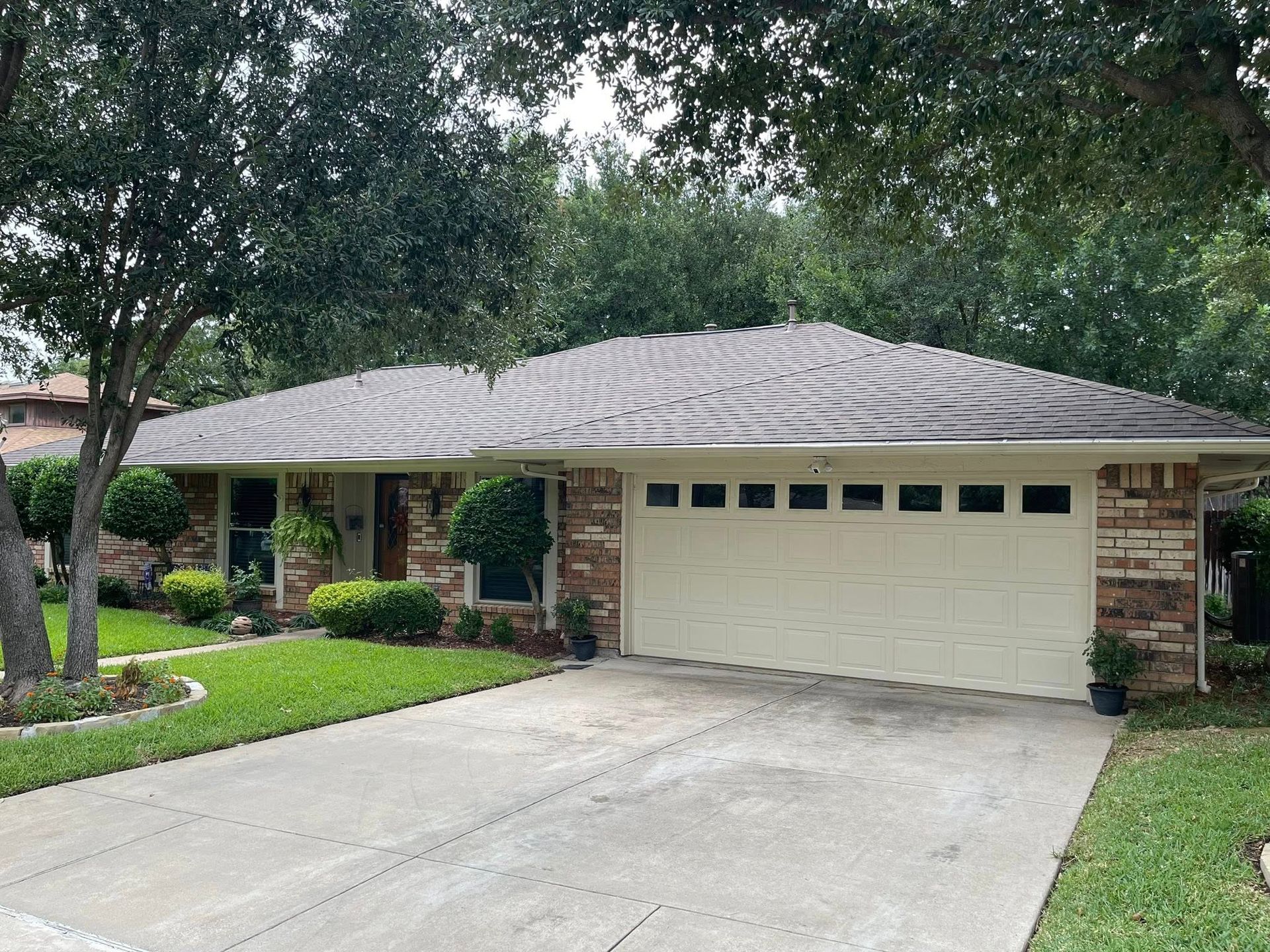 A one-story brick house with a beige garage door and a concrete driveway under leafy trees.