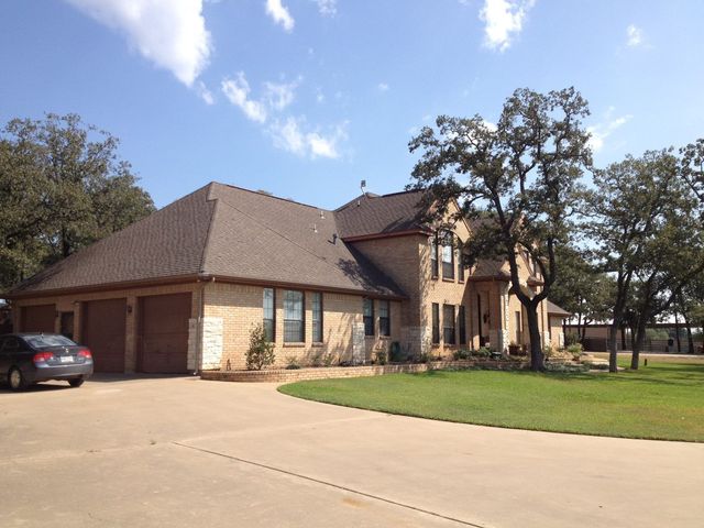 Tan brick house with brown roof, driveway, and trees under a blue sky.