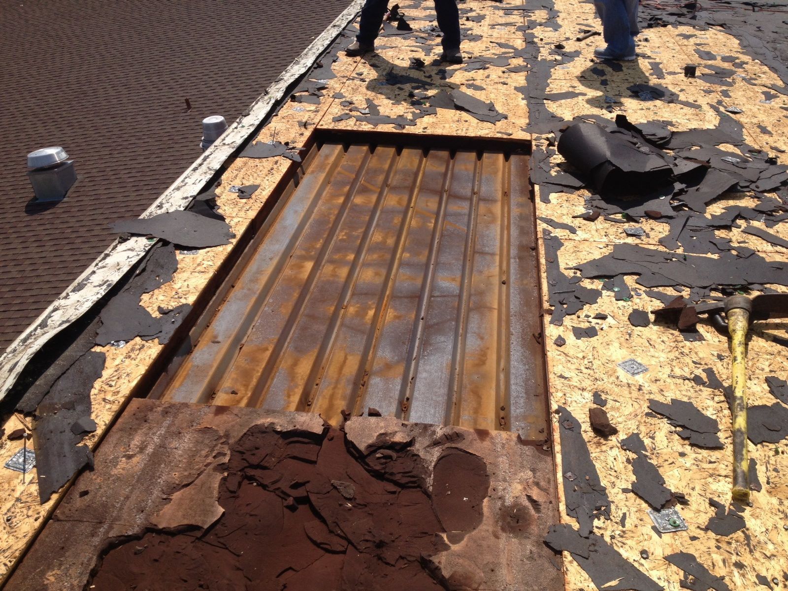Workers removing roofing material, revealing rusty metal decking on a sloped roof.