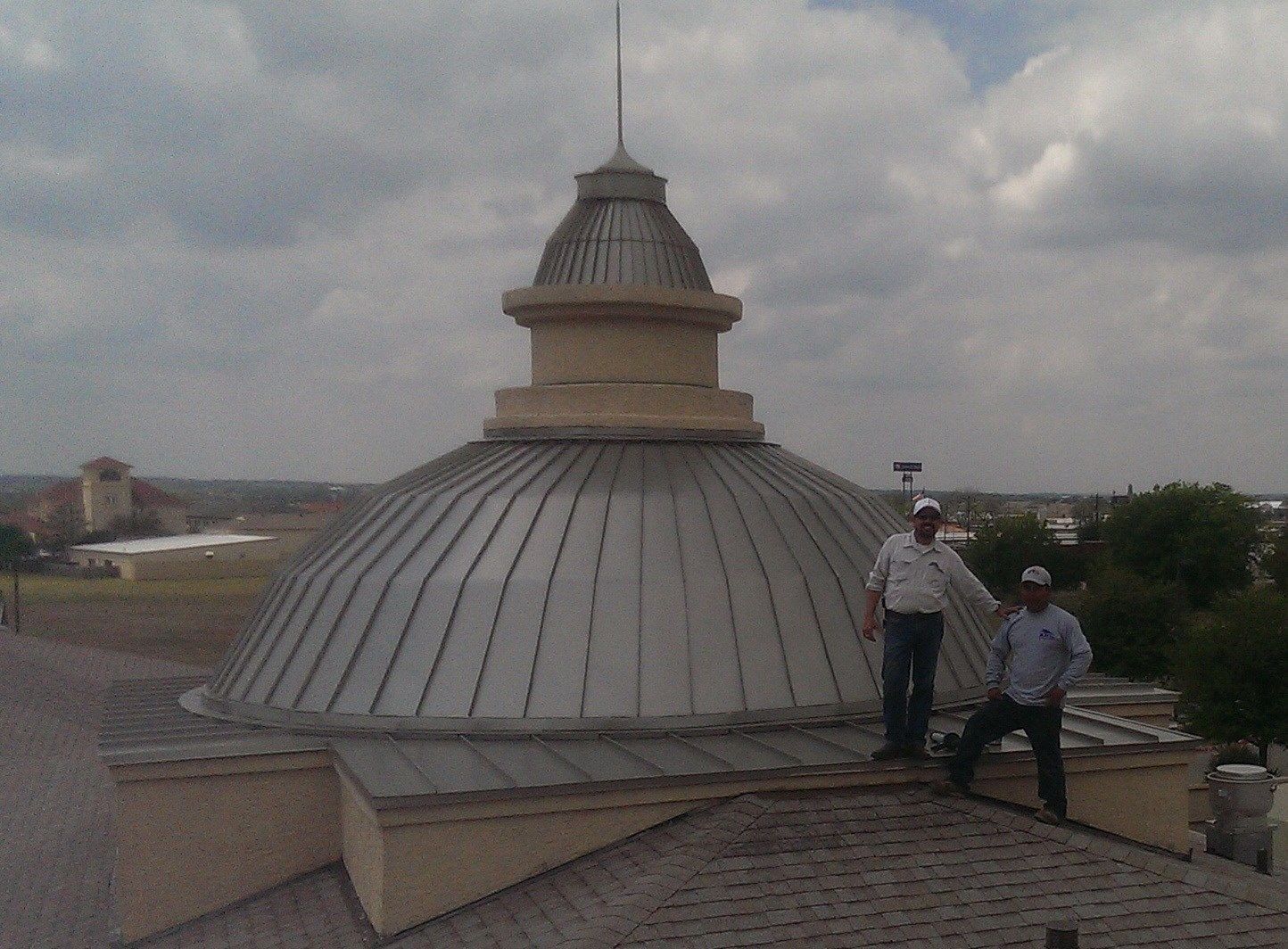 Two men stand on a roof near a large metal dome, under a cloudy sky.