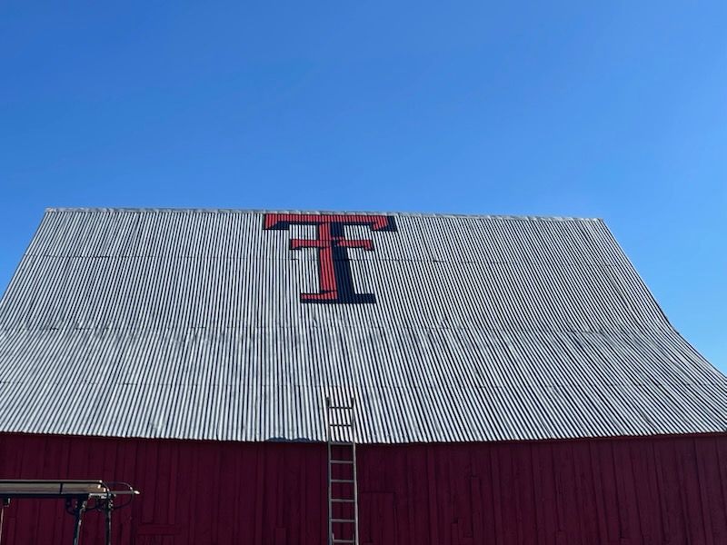 Red barn with a gray metal roof and a large Texas Tech University “T” emblem against a blue sky