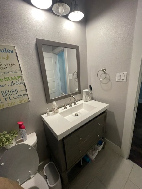 Small bathroom vanity with mirror, sink, wall art, and gray tile floor under bright lights