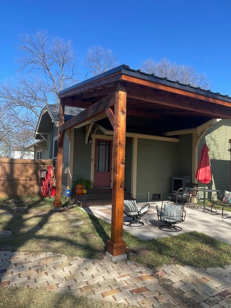 Covered patio with wooden posts, brick pavers, and outdoor chairs beside a house on a sunny day