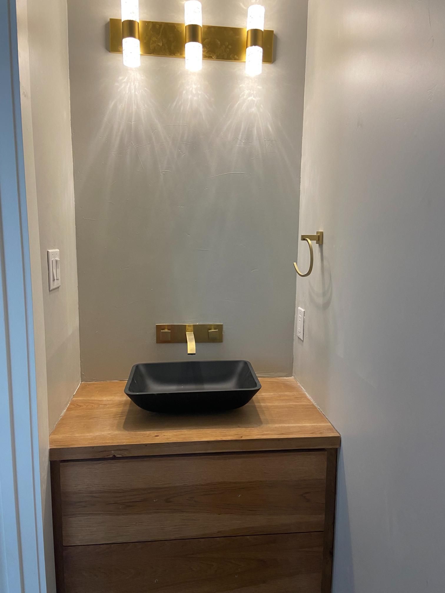 Modern bathroom vanity with wood cabinet, black sink, gold faucet, and wall lights across a gray alcove