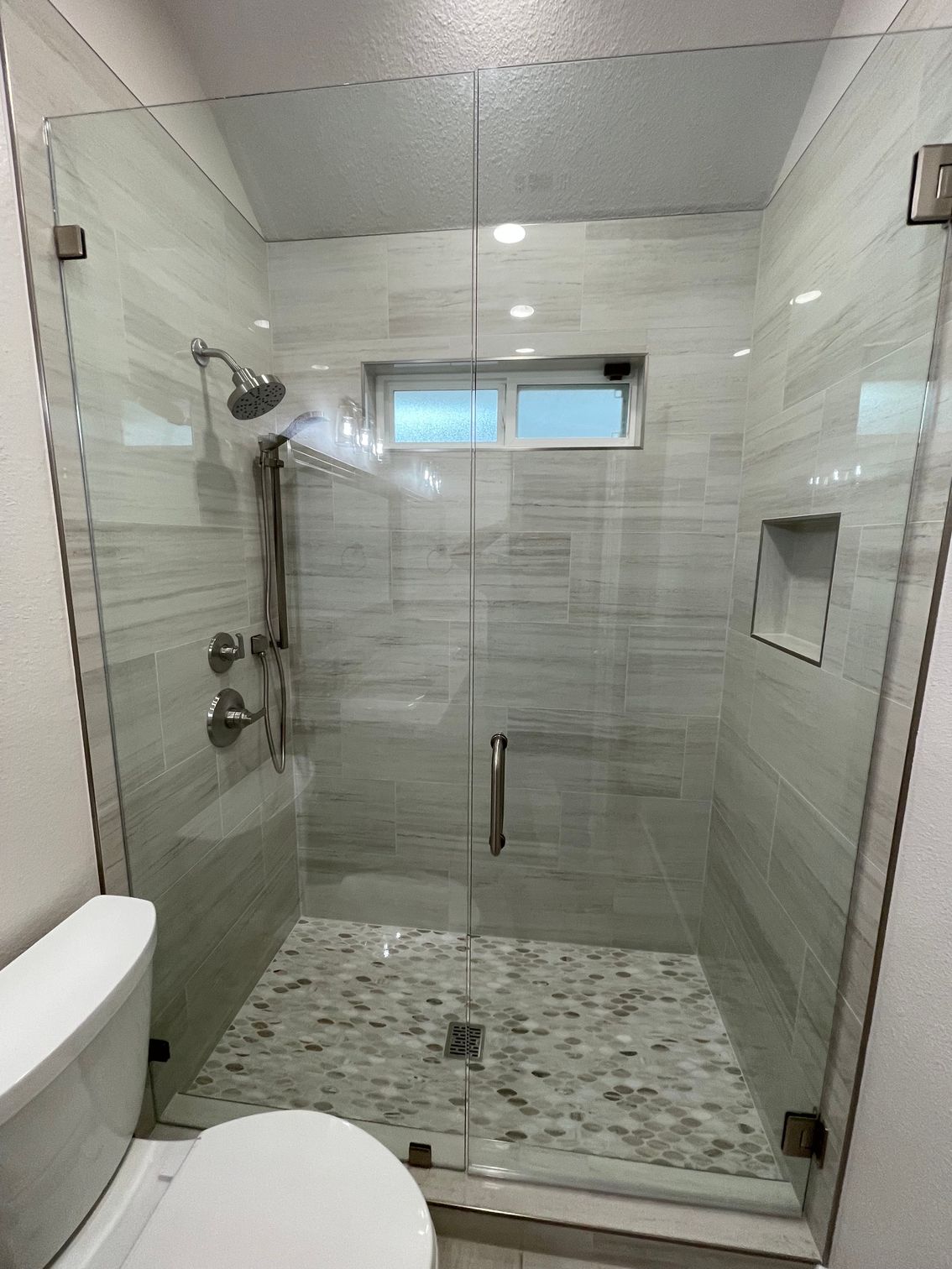 Glass-enclosed tiled shower with rainfall head, built-in shelf, pebble floor, and small window above the door.