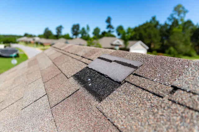 Roofer in red shirt repairing shingles with a hammer on a rooftop.