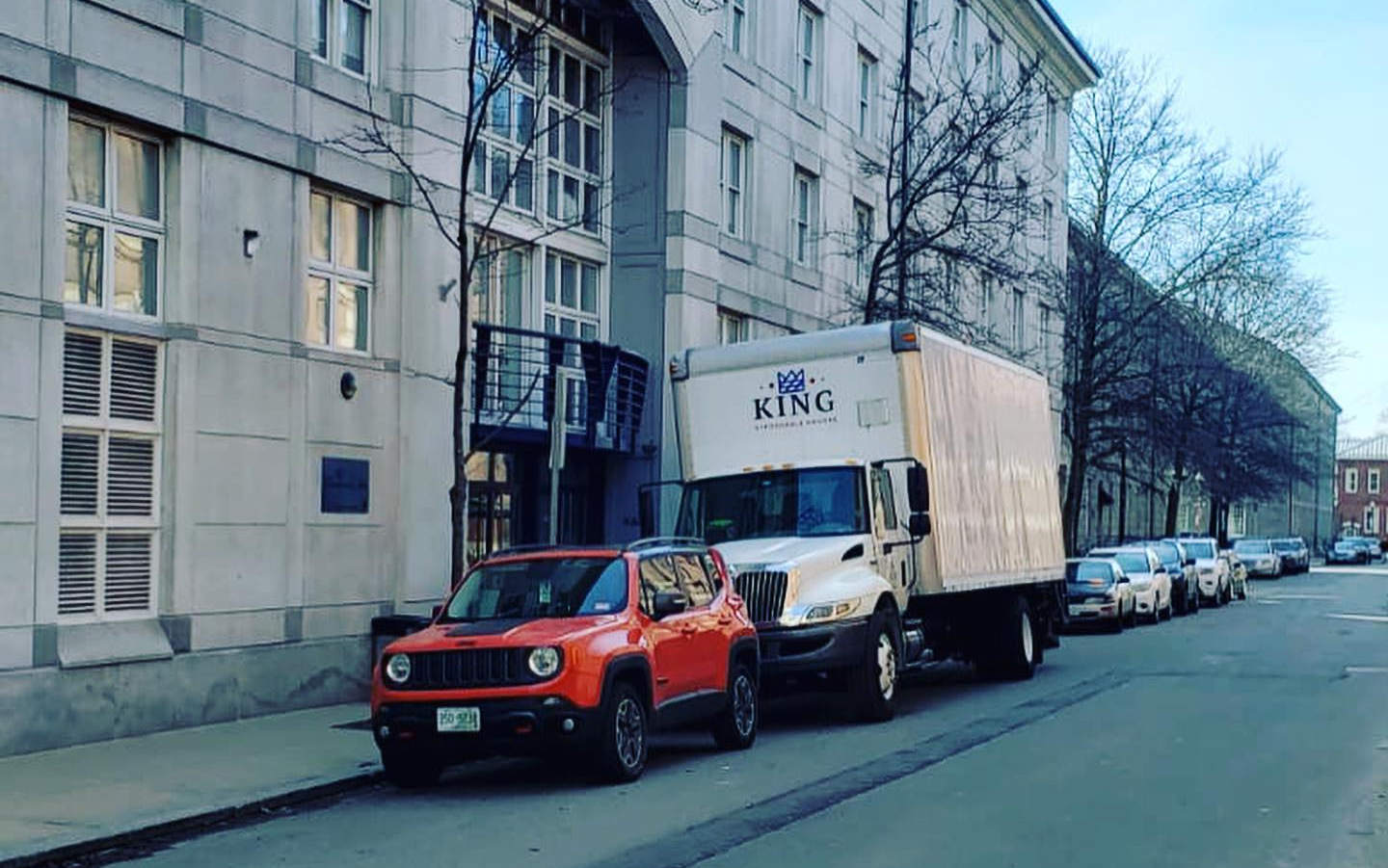 A red jeep and a white truck are parked on the side of the road.