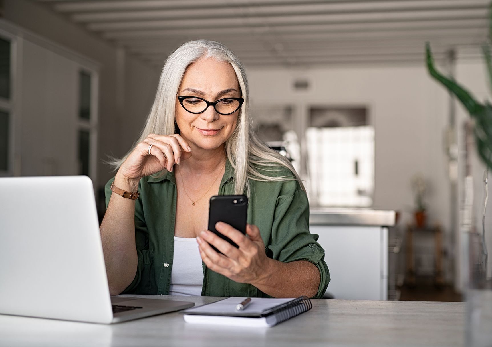 A woman is sitting at a table using a laptop and a cell phone.