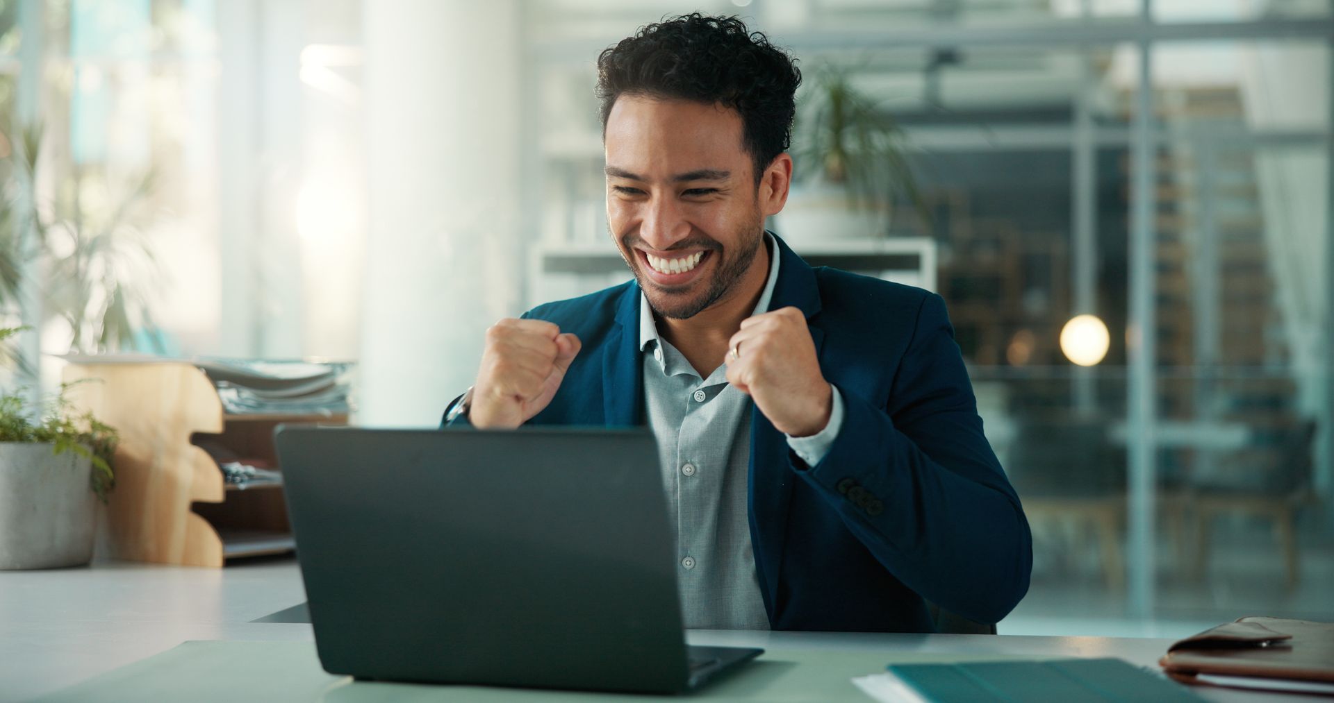 Man in blazer smiles with fists raised at a laptop. Office setting with natural light.