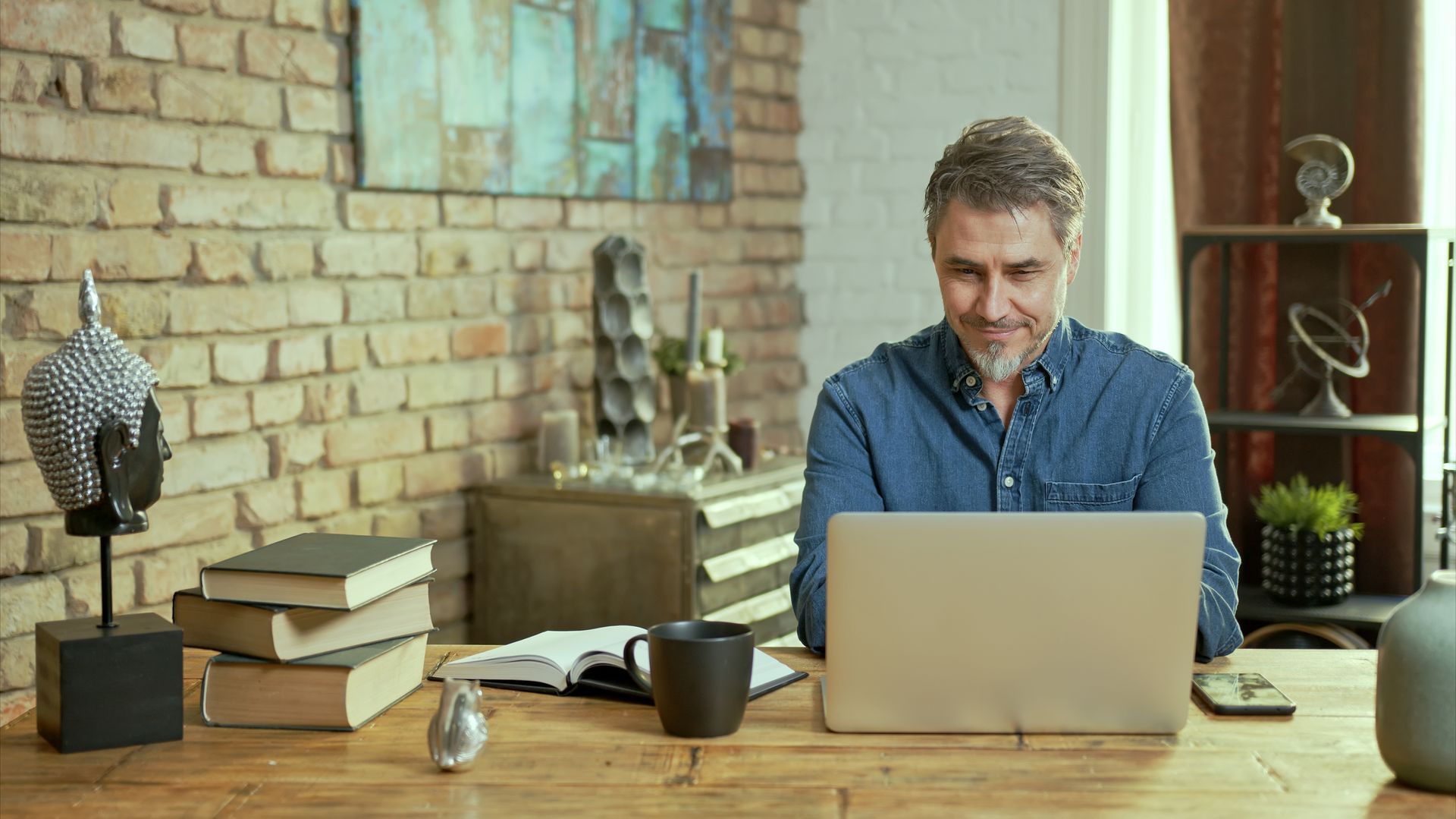 Man smiling at a laptop on a wooden table in a cozy home office with books and decor