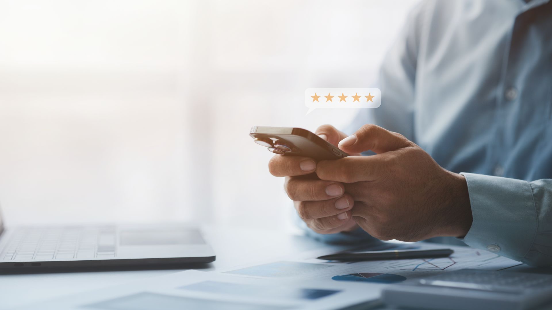 Person using a smartphone at a desk beside a laptop, with a small star rating icon above the phone