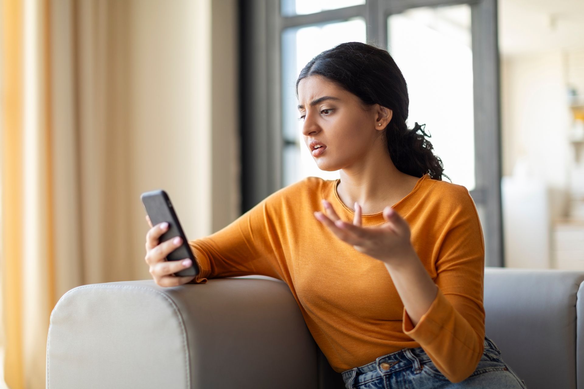 Woman in an orange sweater speaking on a phone while sitting on a couch in a bright room