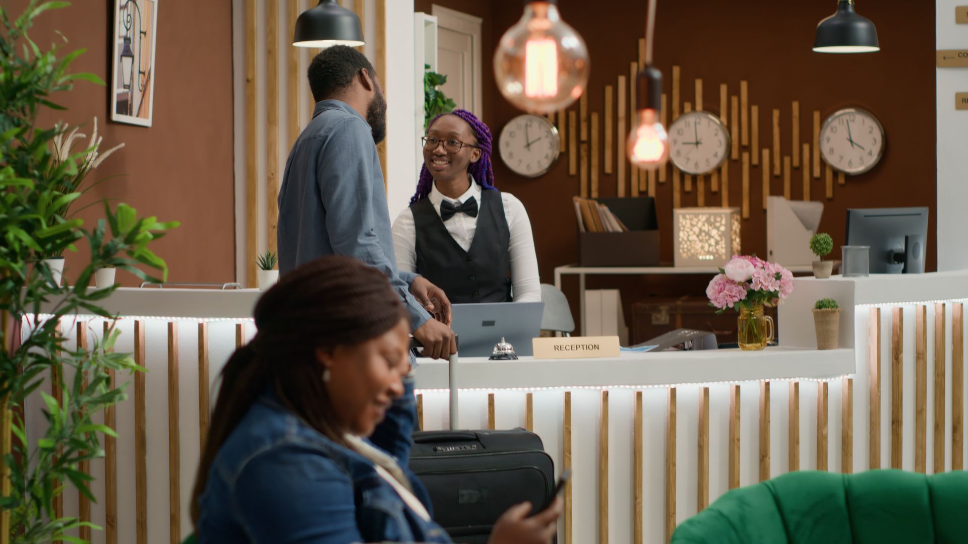 Hotel counter with two staff, hanging lights, clocks, and a seated customer in the foreground.