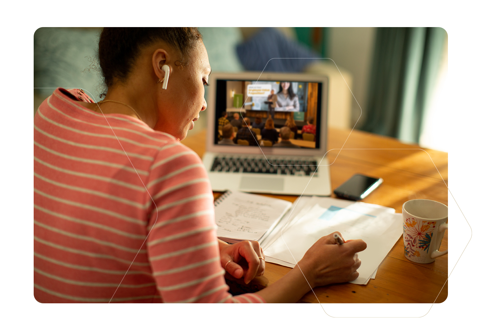 Woman in pink striped shirt attends a video meeting on a laptop at a table, writing notes.
