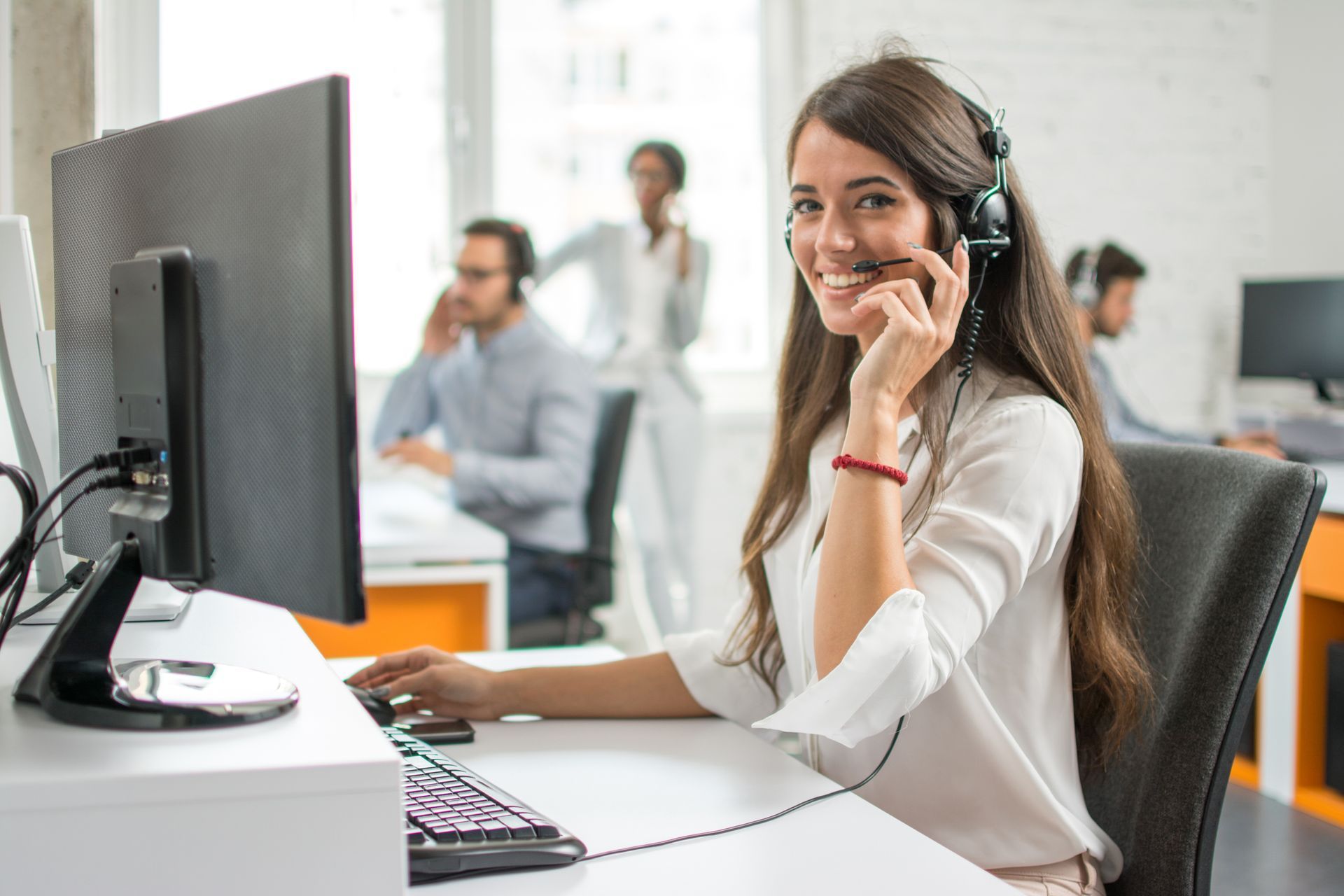 Woman with headset smiles while working at a computer in a bright office; other people also wearing headsets.