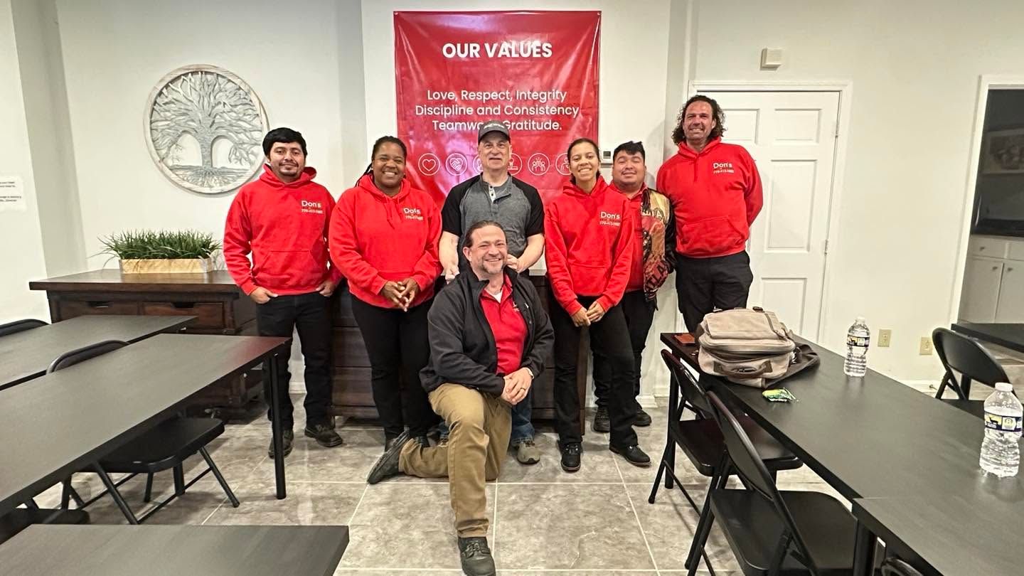 Group posing in a room, five in red jackets behind a seated man with a red banner in the background