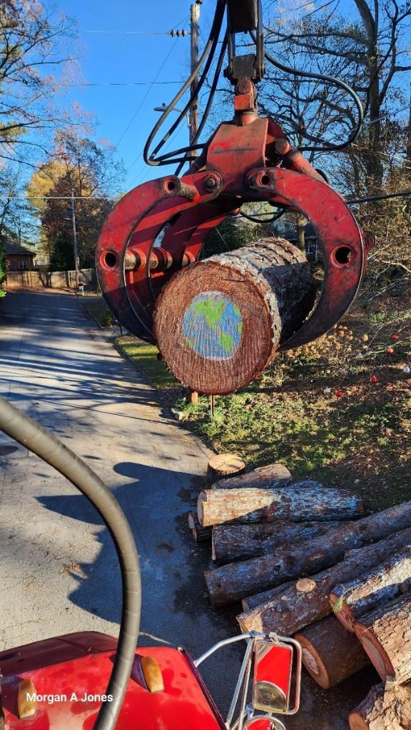 Red logging claw lifting a cut tree trunk beside a roadside woodpile in autumn.
