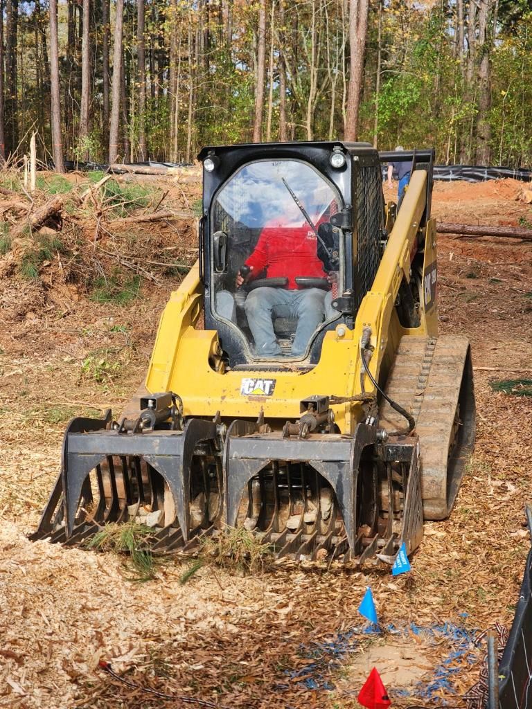 Yellow skid steer with grapple bucket moving leaves on a wooded dirt lot