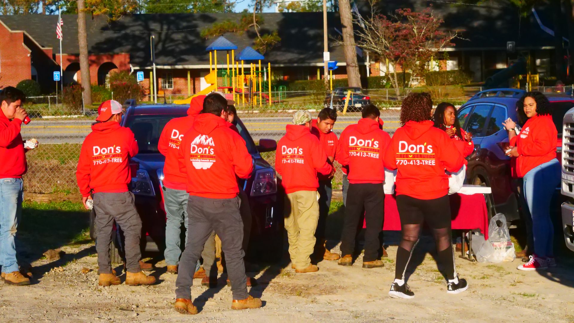 Group of people in red shirts standing outdoors in a park, seen from behind, with trees and cars in the background