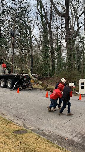 Two workers in red jackets pull a thick cable near a road beside a trailer and orange cones.