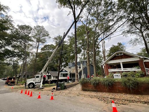 Utility trucks and a bucket lift beside a house under tall trees, with orange cones blocking the road.