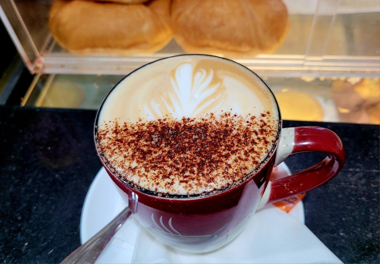 Red cup of latte with foam art and cocoa on a saucer, on a café counter