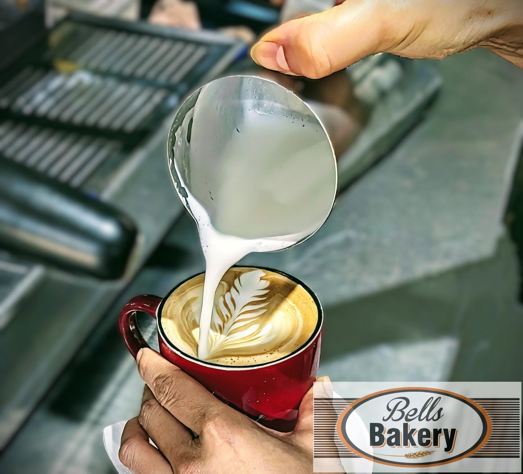 Barista pours steamed milk into a red mug of latte art, with Bell’s Bakery logo visible.