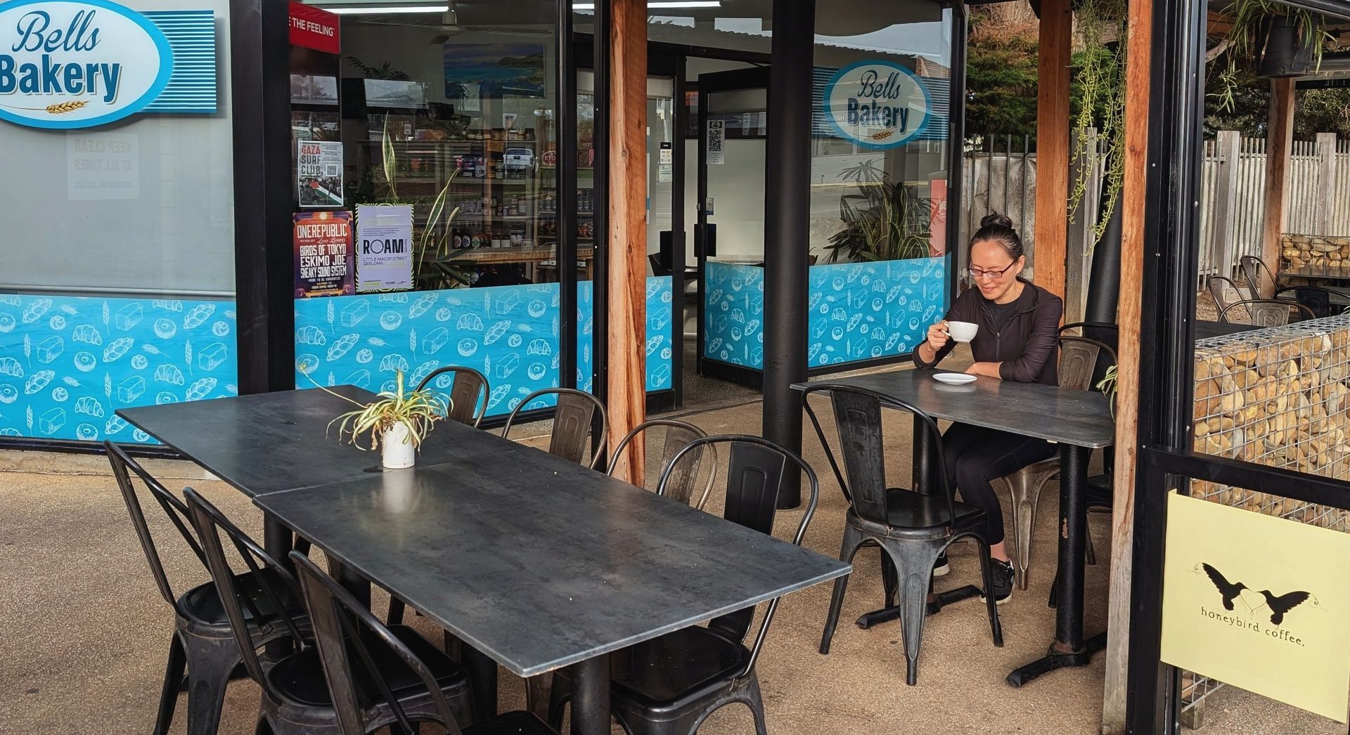 Outdoor bakery seating with black tables and chairs; a person sits by the window on a sunny patio.