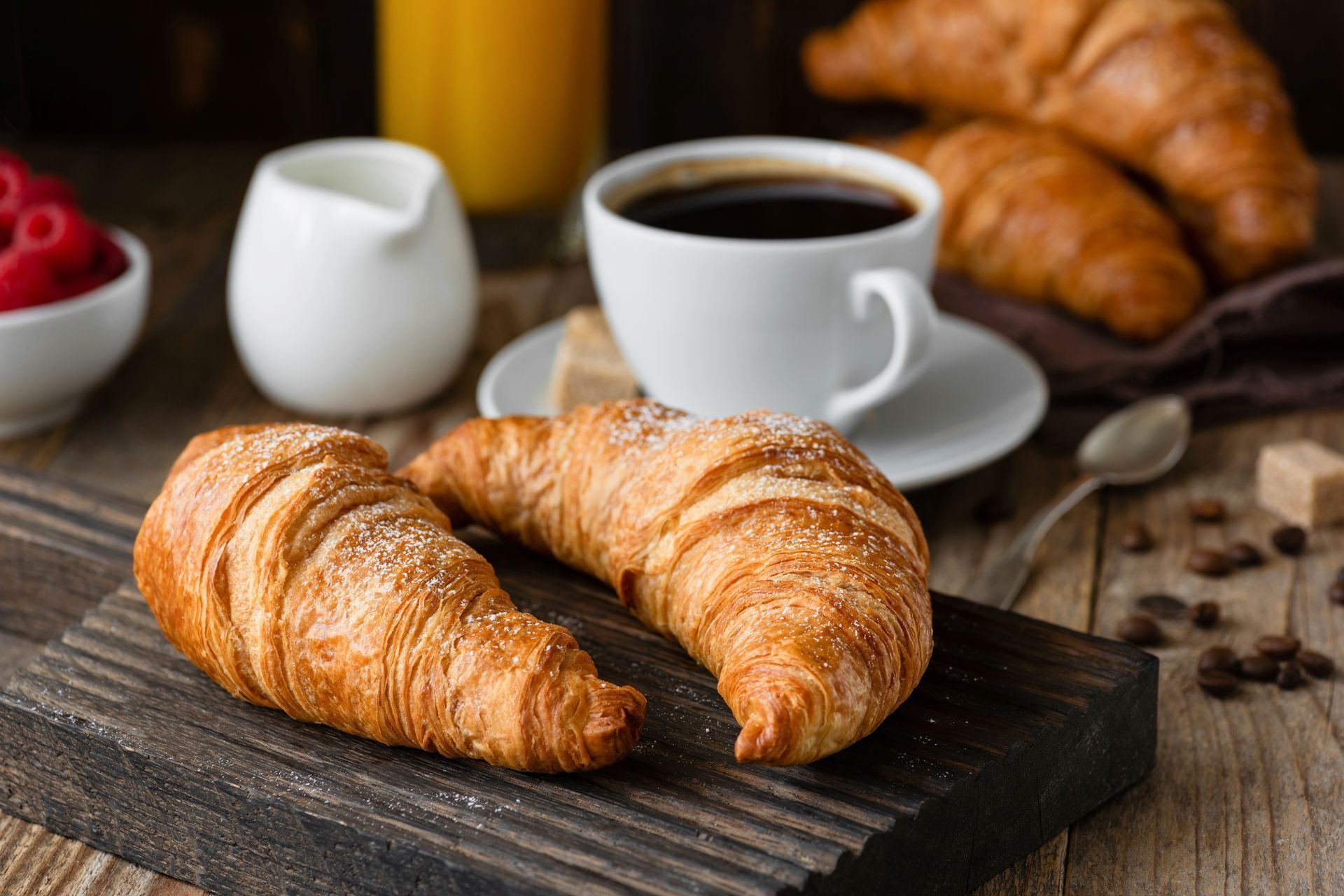 Croissants on a rustic table with coffee cups and raspberries in the background