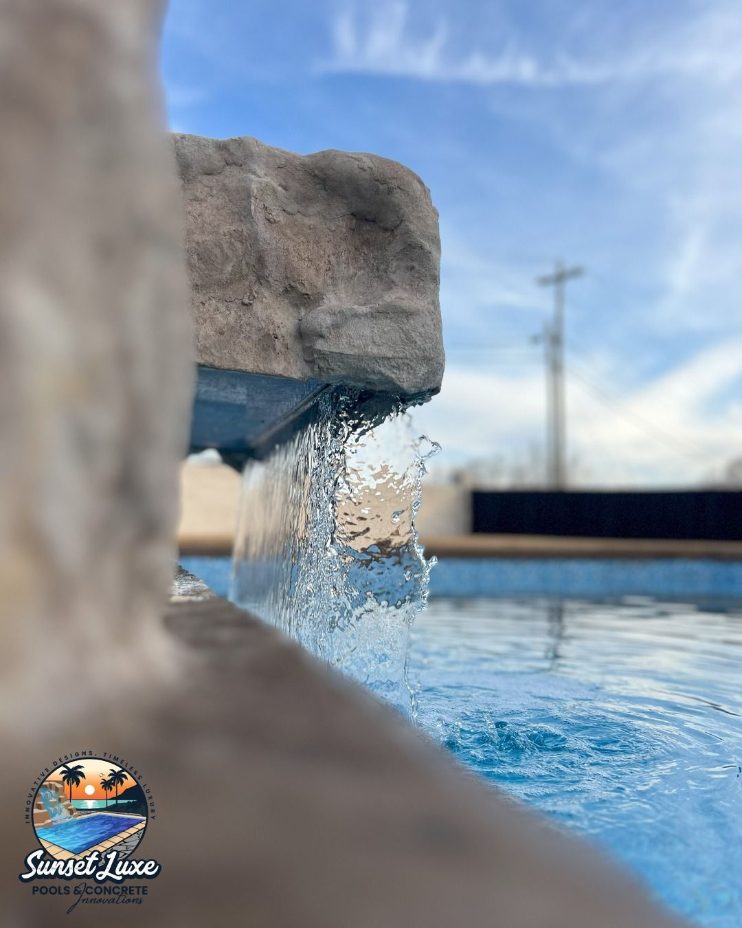 Small waterfall spilling into a blue swimming pool under a cloudy sky.
